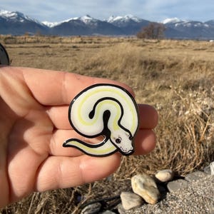 May include: A white and yellow snake pin with a black outline. The pin has a detailed snake head and is held in a hand. The text "No AI used in my Art" is in the upper right corner. The background features mountains and a field.