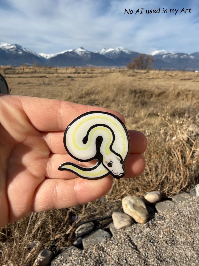 May include: A white and yellow snake pin with a black outline. The pin has a detailed snake head and is held in a hand. The text "No AI used in my Art" is in the upper right corner. The background features mountains and a field.