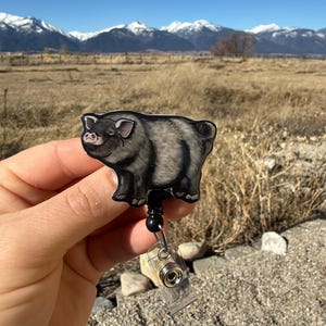 May include: A black and gray pig-shaped retractable badge holder. The pig has a pink snout and ears. The badge holder has a black cord, a black bead, and a clear plastic clip. Text in the image reads: "No AI used in my Art."