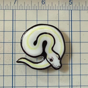 May include: A white and yellow snake-shaped enamel pin with a black outline. The pin has a coiled body and a detailed head with red eyes. The pin is on a grid background.