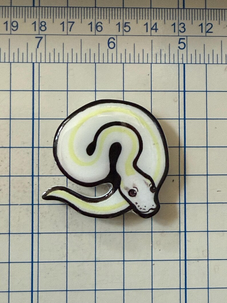 May include: A white and yellow snake-shaped enamel pin with a black outline. The pin has a coiled body and a detailed head with red eyes. The pin is on a grid background.