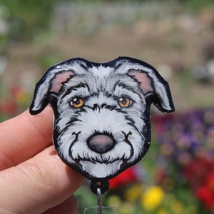 May include: A gray and white dog head with brown eyes, a black nose, and a white beard. The dog is depicted in a cartoon style and is printed on a badge.