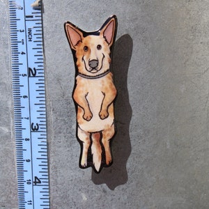 May include: A hand-painted wooden pin of a cartoon Australian Shepherd dog standing on its hind legs. The dog is brown and white with black eyes and a black nose. The pin is about 2 inches tall.