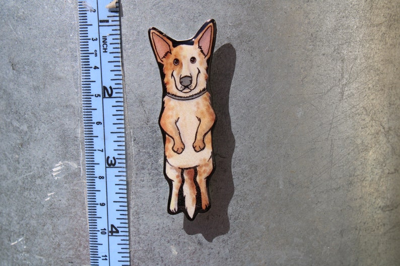 May include: A hand-painted wooden pin of a cartoon Australian Shepherd dog standing on its hind legs. The dog is brown and white with black eyes and a black nose. The pin is about 2 inches tall.