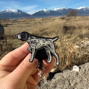 May include: A badge reel featuring a German Shorthaired Pointer dog design. The dog is black and white with a black outline. The reel is black with a clear plastic clip and a retractable cord. The text "No AI used in my Art" is visible in the background.