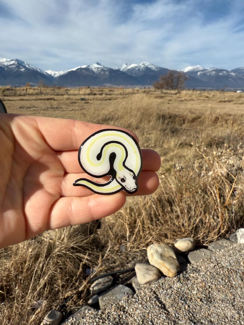 May include: A circular enamel pin featuring a coiled snake design. The snake has a white body with a yellow stripe and black outline. The pin is held in front of a scenic background of mountains and a field.