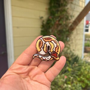 May include: A colorful snake-shaped pin with a detailed design. The pin features a coiled snake with a pattern of yellow, white, and brown. The snake's head is a deep red color. The pin is held in a hand.