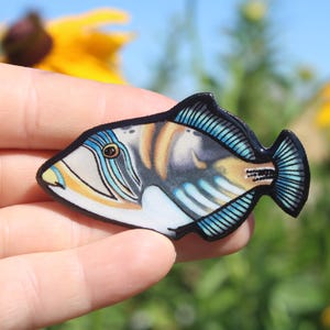 May include: A colorful, detailed illustration of a triggerfish with blue, white, and brown markings. The fish is depicted in a side profile, with its mouth open and its fins spread out. The illustration is printed on a black background.