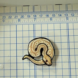 May include: A coiled snake-shaped pin with a black outline. The snake has a tan and cream pattern with brown spots and a red eye. The pin is on a blue and white grid.