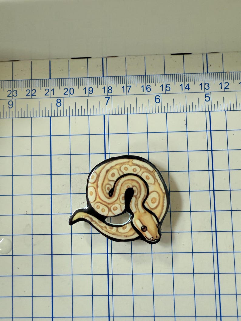 May include: A coiled snake-shaped pin with a black outline. The snake has a tan and cream pattern with brown spots and a red eye. The pin is on a blue and white grid.