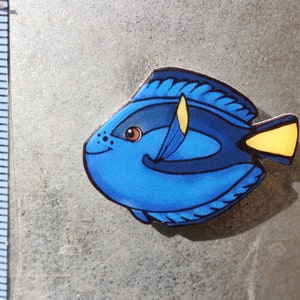 May include: A blue and yellow fish figurine with a black outline. The fish has a large, round body and a small, pointed tail. The fish's eyes are brown and have black pupils. The fish is painted with a blue body, a yellow tail, and a black outline.