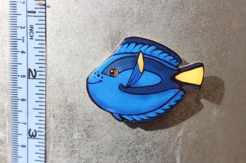 May include: A blue and yellow fish figurine with a black outline. The fish has a large, round body and a small, pointed tail. The fish's eyes are brown and have black pupils. The fish is painted with a blue body, a yellow tail, and a black outline.