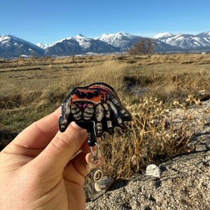 May include: A badge reel featuring a colorful spider design. The spider has a black body, orange eyes, and white and black striped legs. The reel has a black retractable cord and a clear plastic clip.