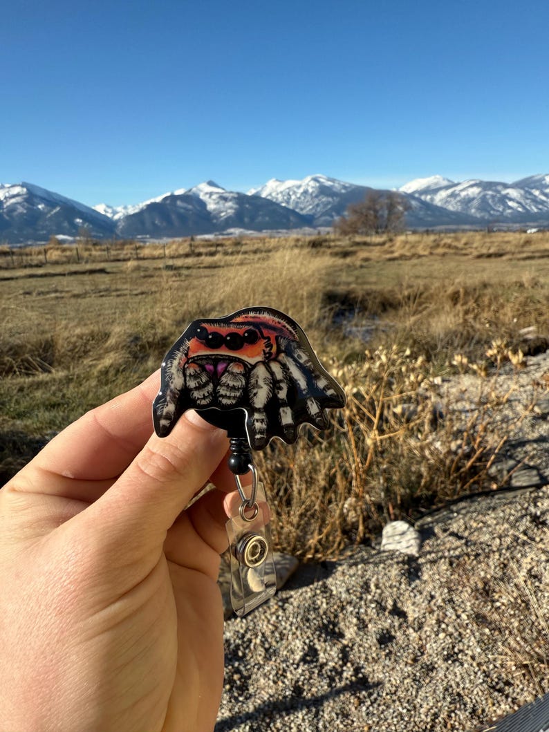 May include: A badge reel featuring a colorful spider design. The spider has a black body, orange eyes, and white and black striped legs. The reel has a black retractable cord and a clear plastic clip.