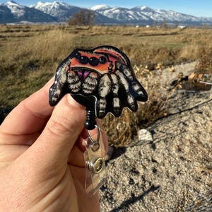 May include: A black and white spider badge reel with a retractable cord and a clear plastic ID holder. The spider has orange, pink, and black details. The text "No AI used in my Art" is visible in the background.
