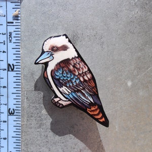 Op de afbeelding: Een houten uitsnijding van een kookaburra met blauwe, bruine en witte veren. De vogel zit op een tak en heeft een lange, puntige snavel.