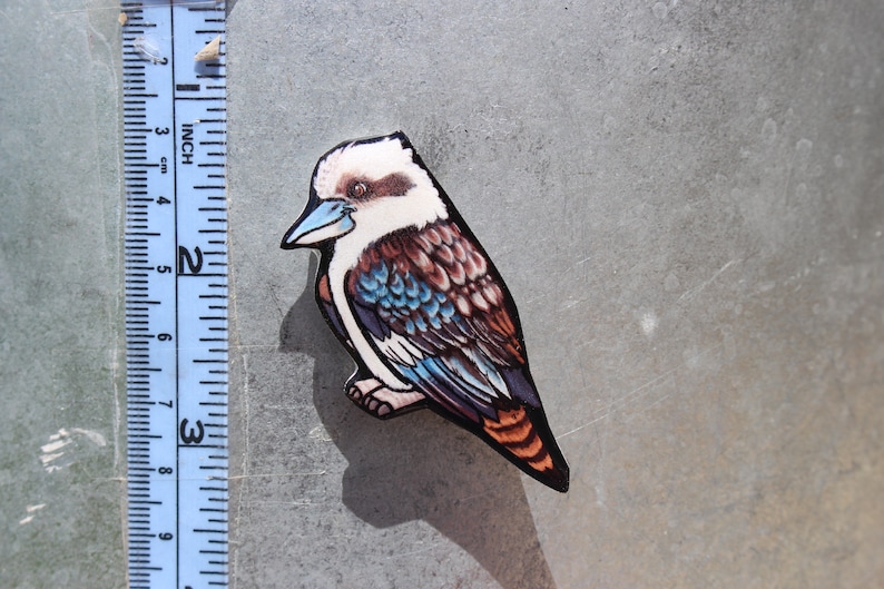Op de afbeelding: Een houten uitsnijding van een kookaburra met blauwe, bruine en witte veren. De vogel zit op een tak en heeft een lange, puntige snavel.