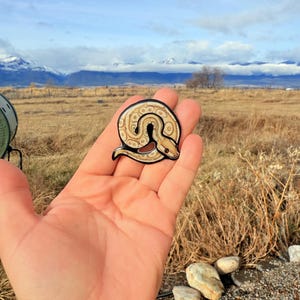 May include: A circular snake-shaped pin with a cream and brown pattern, outlined in black. The pin is held in a hand against a backdrop of a field, mountains, and a cloudy sky. The pin is a decorative accessory.