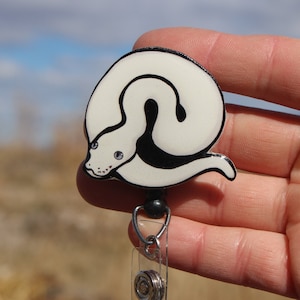 May include: White and black snake design on a retractable badge holder. The snake is curled up with its head facing the viewer. The badge holder is made of metal and has a clear plastic strap.