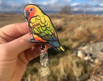 May include: A colorful sun conure bird badge reel with a black retractable cord and a clear plastic badge holder. The bird is depicted in a realistic style with bright yellow, orange, red, green, and blue feathers.