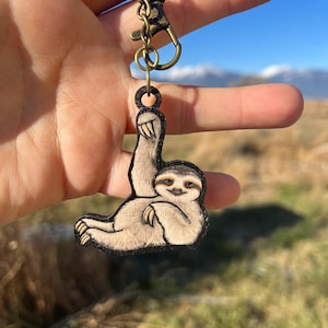 May include: A wooden keychain featuring a sloth with a brown and black painted design. The sloth is hanging upside down with its arms outstretched. The keychain is attached to a bronze metal chain and clasp.