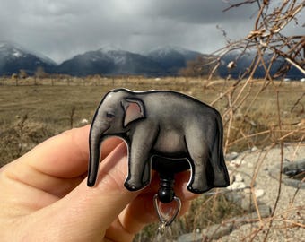 May include: A gray elephant cartoon badge reel with a black retractable cord and a clear plastic clip. The badge reel is held in a hand against a background of a mountain range and a cloudy sky.