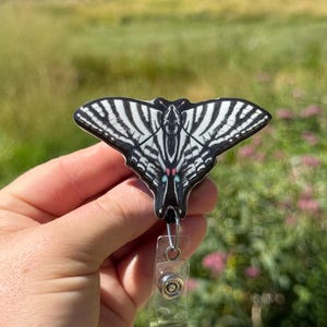 May include: A black and white butterfly badge reel with a retractable cord and a clear plastic clip. The butterfly design features intricate wing patterns with red and pink accents. The badge reel is held against a blurred outdoor background.