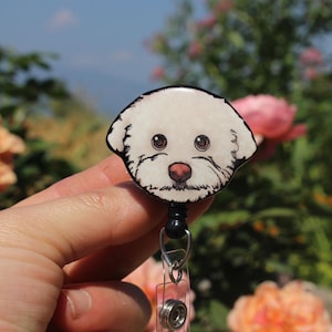 May include: A white dog face badge reel with a black retractable cord. The dog has black eyes and a pink nose.