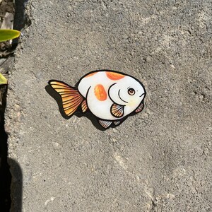 May include: A white and orange goldfish sticker with black outlines. The goldfish has a round body and a small tail. The sticker is on a gray concrete surface.