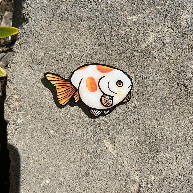 May include: A white and orange goldfish sticker with black outlines. The goldfish has a round body and a small tail. The sticker is on a gray concrete surface.