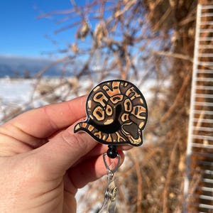 May include: A brown and black snake curled up on a white background. The snake is attached to a clear plastic retractable badge holder with a black clip.