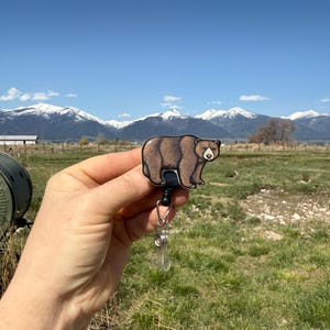 May include: A brown bear cartoon character on a retractable badge holder. The bear is facing forward with a neutral expression. The badge holder is black and silver. The background is a blurry image of a mountain range and a grassy field.