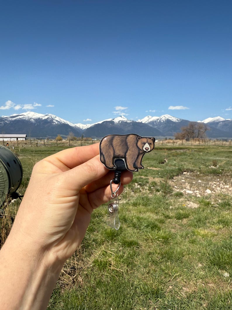 May include: A brown bear cartoon character on a retractable badge holder. The bear is facing forward with a neutral expression. The badge holder is black and silver. The background is a blurry image of a mountain range and a grassy field.