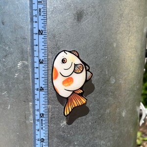 May include: A sticker of a cartoon fish with orange and white scales. The fish is about 2 inches long and has a smiling face.