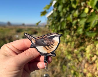 May include: A badge reel featuring a detailed illustration of a small bird with brown, tan, and white feathers. The bird is attached to a black retractable reel with a clear plastic strap and a metal clip. The background is a natural outdoor scene.