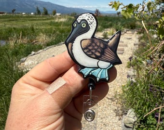 May include: A blue-footed booby bird badge reel with a black retractable cord and a clear plastic holder. The bird has a white head with black spots, a brown body, and light blue feet. The badge reel is held in front of a natural outdoor background.