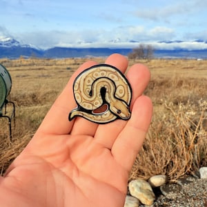 May include: A circular, glossy sticker featuring a coiled snake design. The snake has a tan and brown pattern with a black outline. The sticker is held in a hand, with the text "No AI used in my Art" visible in the background.