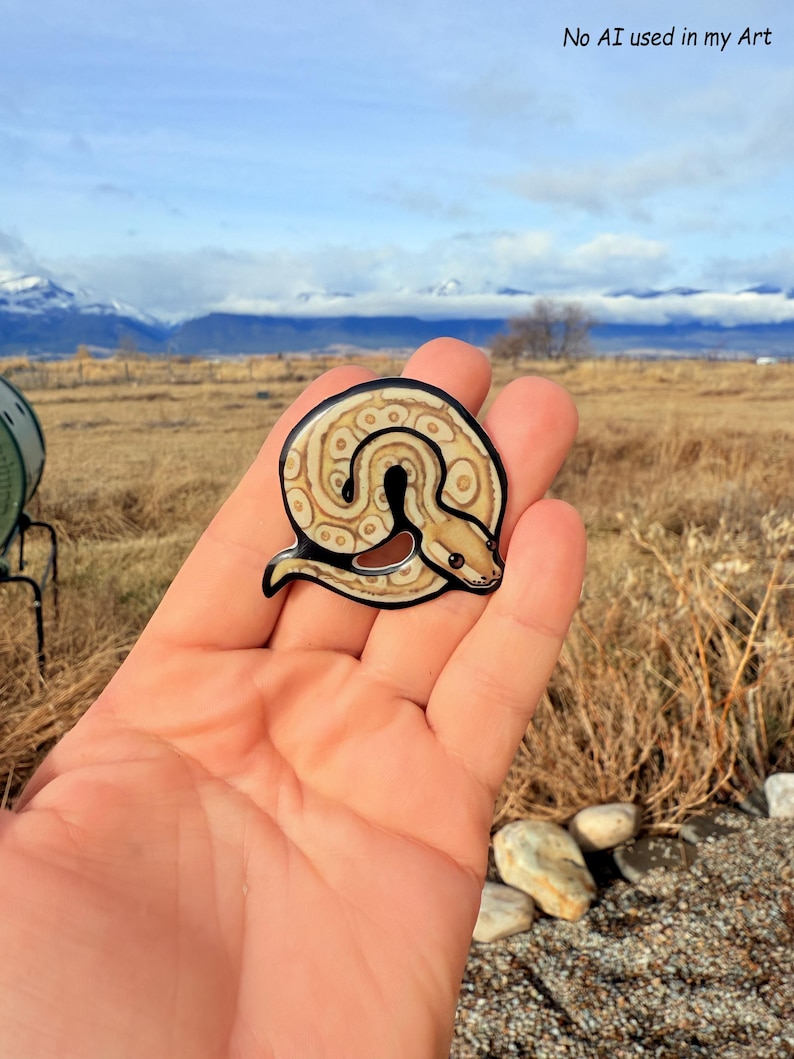 May include: A circular, glossy sticker featuring a coiled snake design. The snake has a tan and brown pattern with a black outline. The sticker is held in a hand, with the text "No AI used in my Art" visible in the background.