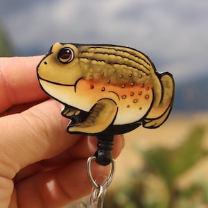 May include: A brown and yellow frog with orange spots is attached to a retractable badge reel. The frog is cartoon-like and has a 3D effect.
