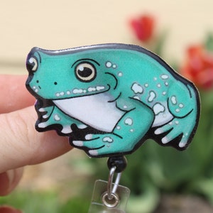 May include: A teal green frog with white spots, sitting on a clear plastic badge holder with a black swivel clip. The frog has a white belly and black eyes.