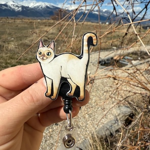 May include: A white and orange cat cartoon badge reel with a black retractable cord and a clear plastic clip. The cat is looking to the left and has green eyes.