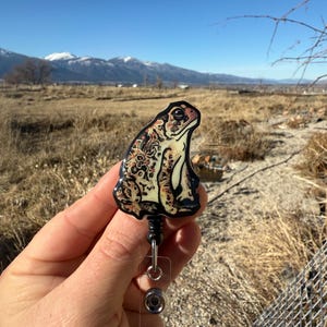 May include: A badge reel featuring a detailed illustration of a toad with a mottled pattern in shades of brown, tan, and cream. The badge reel has a black retractable cord and a clear plastic holder. The text "No AI used in my Art" is visible.