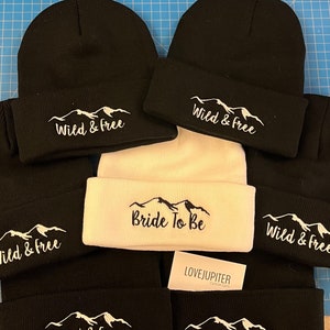 Puede incluir: Seis gorros de punto negros con bordado blanco de monta&ntilde;a y texto. Un gorro es blanco con bordado negro de monta&ntilde;a y texto. El texto en los gorros negros dice "Wild & Free". El texto en el gorro blanco dice "Bride To Be".