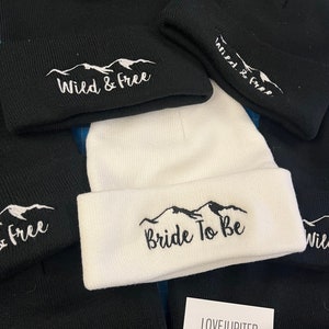 Puede incluir: Una colecci&oacute;n de gorros de punto negros y blancos con monta&ntilde;as bordadas y texto. Los gorros presentan el texto "Wild & Free" y "Bride To Be".