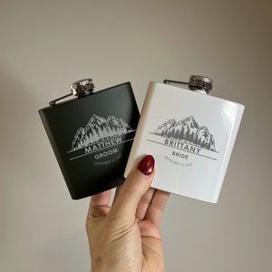 May include: Two black and white metal hip flasks with a mountain design and the words "Groom" and "Bride" with the names "Matthew" and "Brittany" and the date "September 10, 2022".