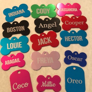 May include: An assortment of personalised dog tags in various colours and shapes, including bone and rectangular shapes. The tags are engraved with names such as Cody, Cassandra, Boston, Angel, Cooper, Louie, Jack, Hector, Abagail, Freya, Oscar, Coco, Millie, and Oreo.