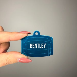 May include: A teal pet tag in the shape of a barrel, with the name "BENTLEY" in white block letters. The tag has a hole at the top for attachment and is held against a gray background.
