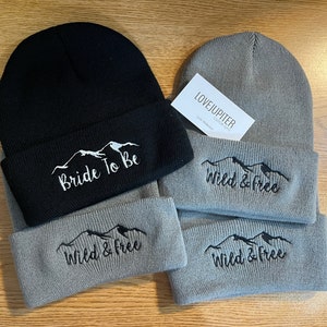 Puede incluir: Dos gorros de punto, uno negro y otro gris, con monta&ntilde;as bordadas y texto. El gorro negro dice "Bride To Be" y el gorro gris dice "Wild & Free".