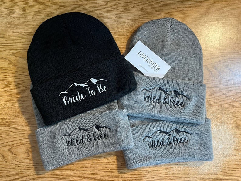 Puede incluir: Dos gorros de punto, uno negro y otro gris, con monta&ntilde;as bordadas y texto. El gorro negro dice "Bride To Be" y el gorro gris dice "Wild & Free".