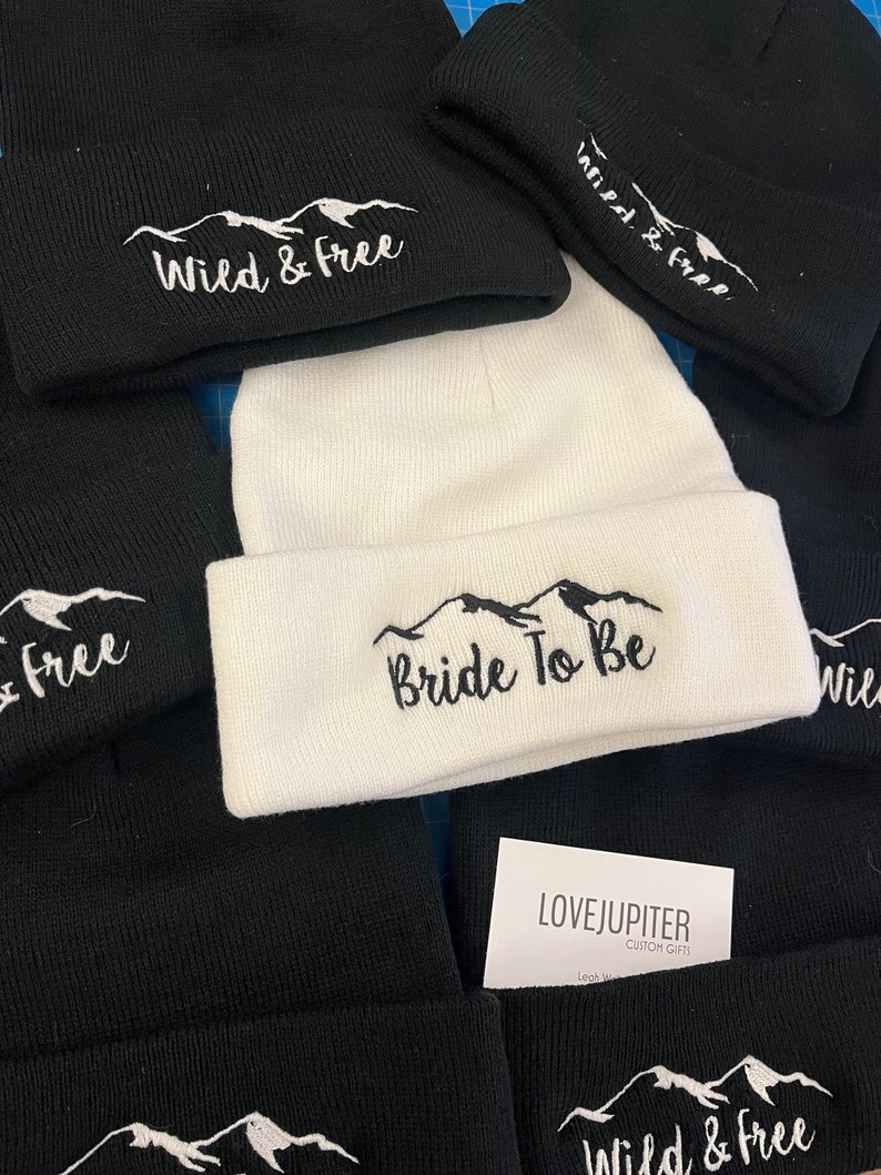 Puede incluir: Una colecci&oacute;n de gorros de punto negros y blancos con monta&ntilde;as bordadas y texto. Los gorros presentan el texto "Wild & Free" y "Bride To Be".